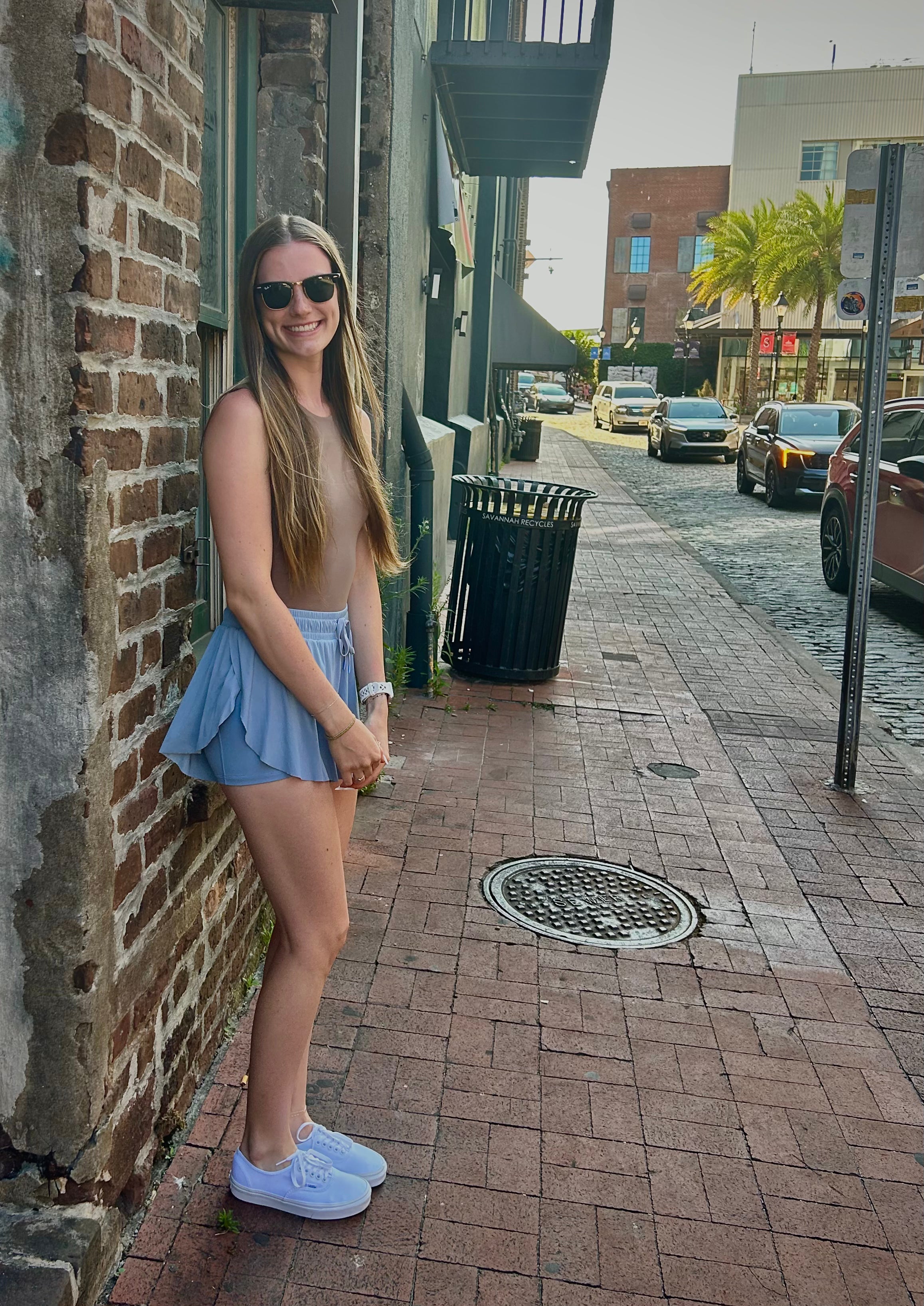 Woman in blue shorts, brown body suit, and white sneakers standing on a sidewalk next to a brick wall.